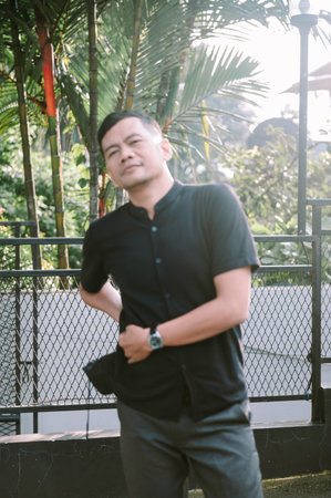A confident looking man in casual wear poses outdoors next to a metal fence with lush tropical plants in the background.の写真素材
