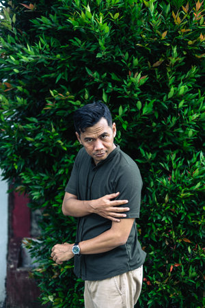 A man poses near a leafy green hedge with a watch.