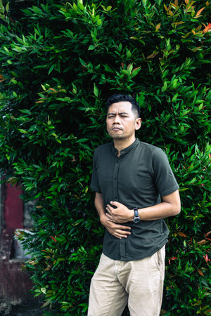 A man is suffering from a stomach ache, holding his stomach while standing in front of a lush green...
