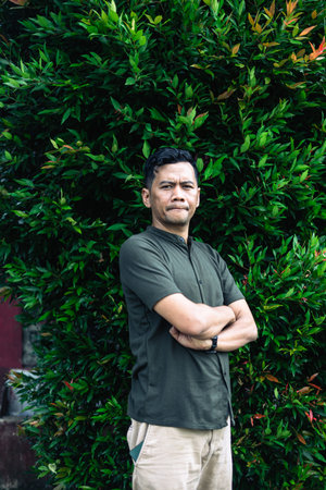 A portrait of a man standing confidently with arms crossed in front of a green leafy bush outdoors.