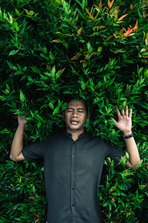 An adult male lying in a dense green leafy shrubbery, surrounded by green foliage, integrating with...