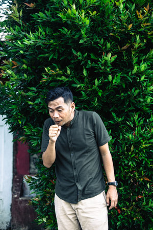 A man is coughing into his hand while standing outside near a lush green bush, symptom of sickness.