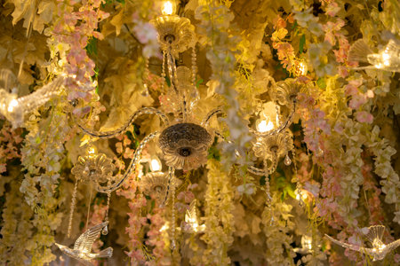Beautiful ceiling decoration featuring a glowing crystal chandelier, warm lights, and elegant hanging pastel flowers, creating a luxurious and festive ambiance for a wedding or special event.の写真素材