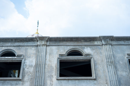 A weathered concrete mosque facade features arched and rectangular windows, classical columns, and a dome topped with a golden crescent and star finial under a bright blue sky.の写真素材