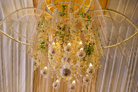 A magnificent crystal chandelier brightly illuminates a beautifully decorated ceiling with cascading white artificial flowers, green leaves, and warm fairy lights, perfect for an elegant event or weddの写真素材