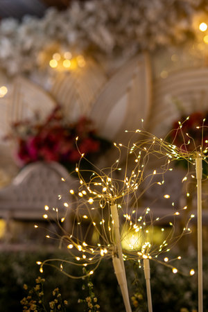 Close-up of decorative warm LED fairy lights on white poles, with a beautifully blurred background featuring elegant event decor, soft fabric, and floral arrangements.の写真素材