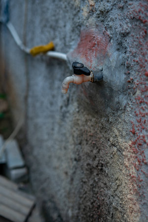 A close-up of an old, off-white plastic water tap with a black handle, slowly dripping water against a rough, grey concrete wall with reddish discoloration.の写真素材