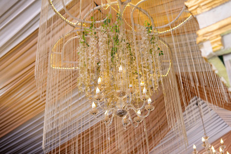 An elegant crystal chandelier adorned with delicate white hanging flowers and shimmering decorative strings, set against a background of softly draped golden and white fabric.の写真素材