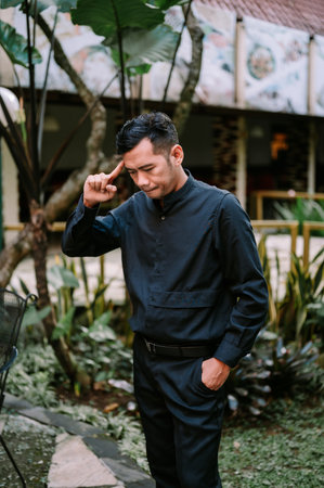 A young adult Asian man in a black band collar shirt and black pants stands outdoors, hand on his head, looking down thoughtfully in a green garden environment.の写真素材