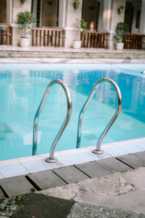 Stainless steel handrails lead into a bright, empty swimming pool with clear turquoise water. The pool edge features white and blue tiles, with blurred resort-style buildings and pottedの写真素材