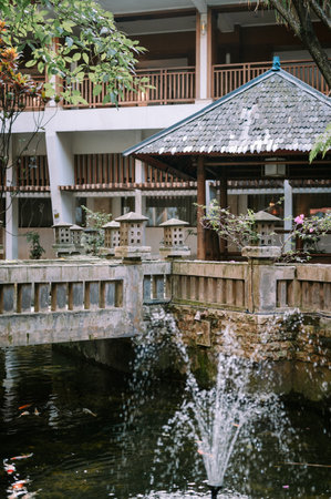 A tranquil outdoor scene featuring a stone bridge, a traditional gazebo with a scaled roof, and a pond with a fountain and colorful koi fish, surrounded by lushの写真素材