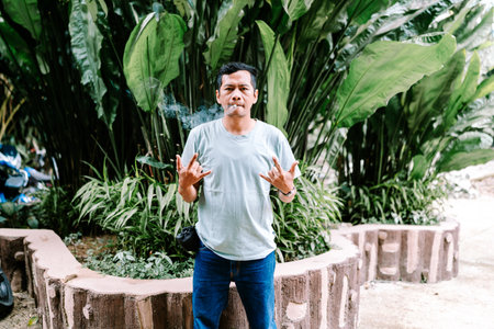 A man stands outdoors, smoking a cigarette and making a rock 'n' roll hand gesture. The lush green foliage provides a natural, tropical background for his expressive pose.の写真素材