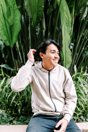 A cheerful young Asian man sits comfortably outdoors, surrounded by vibrant green foliage, expressing happiness and relaxation in a casual moment.の写真素材