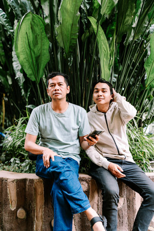 Two men are sitting casually on an outdoor wall, surrounded by vibrant green tropical plants. One man looks at the camera, while the other checks his phone in a relaxed garden setting.の写真素材