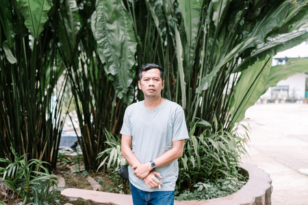 A serious adult Asian man with a calm expression stands against a lush background of tropical plants...