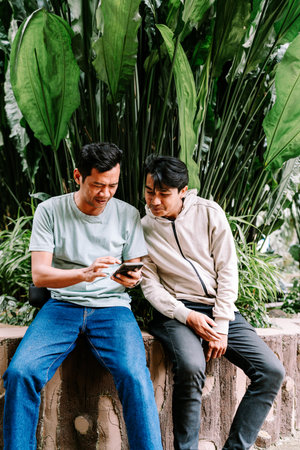 Two young Asian men are sitting outdoors, focused on a smartphone, sharing content or browsing together in a lush tropical setting with vibrant green plants in the background.の写真素材