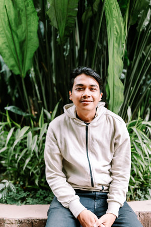 A cheerful young Asian man sits comfortably in a vibrant green garden, smiling genuinely at the camera. He wears a casual hoodie, embodying a relaxed and approachable mood in nature.の写真素材