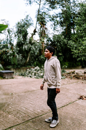A young Asian man walks thoughtfully through a lush green outdoor environment. Dressed casually in a beige hoodie and dark pants, he observes his surroundings amidst tropical foliage and trees.の写真素材