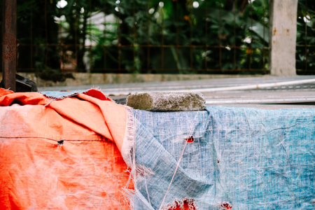 Close-up of heavily worn blue and orange tarpaulins, held down by a rough concrete block. The tattered fabrics display rips and faded colors, creating a gritty textured background reflecting urbanの写真素材