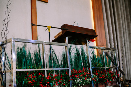 A wooden podium with a microphone stands ready for a speaker in a formal church or ceremonial hall, adorned with vibrant red and green artificial floral arrangements and a sleek metal railing.の写真素材