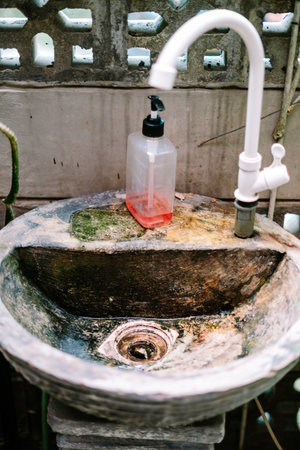 A weathered, handmade concrete sink with a white plastic faucet and a clear soap dispenser bottle, set against a perforated concrete wall in an outdoor setting. It conveys basic hygiene and rusticの写真素材