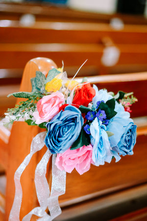 A vibrant artificial flower bouquet with pink, blue, red, and yellow roses beautifully adorns a wooden church pew, perfect for wedding or event decor.の写真素材