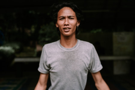A confident young Asian man with dark hair looks directly at the camera in a casual portrait. He wears a simple grey t-shirt, set against a softly blurred background.の写真素材