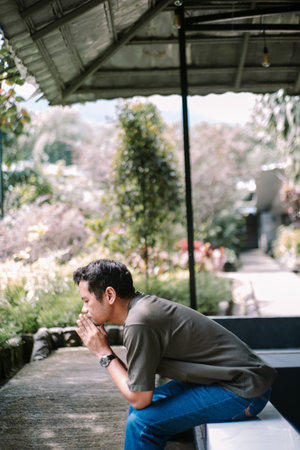 A young man sits quietly outdoors, deep in thought with his hands clasped together. The serene natural background suggests a moment of contemplation or peace.の写真素材