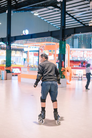 A man with protective gear enjoys rollerblading in a lively indoor leisure center. He moves gracefully on inline skates past colorful kiosks under bright lights.の写真素材
