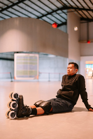 A young man wearing inline skates and protective gear sits on the floor, taking a break from his sport. He is resting after an active session in a modern indoor facility.の写真素材