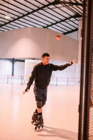 An adult man inline skating on a smooth indoor rink, reaching to catch a small red ball in the air....