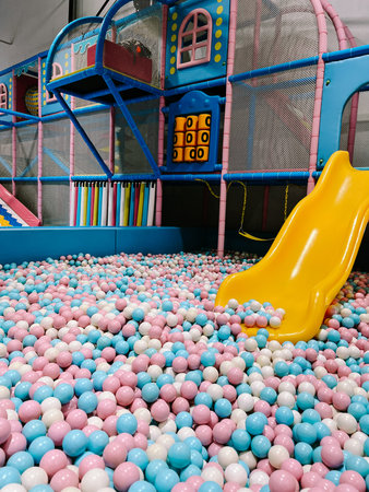 A vibrant indoor playground features a large ball pit filled with soft pink, blue, and white plastic...