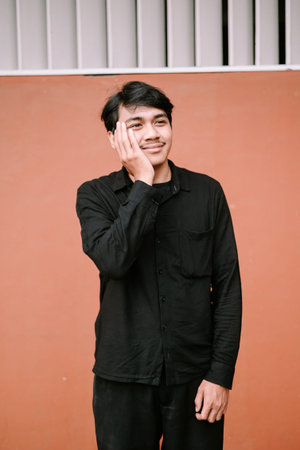 A young Asian man in a black long-sleeved shirt poses against an orange wall, looking thoughtfully...