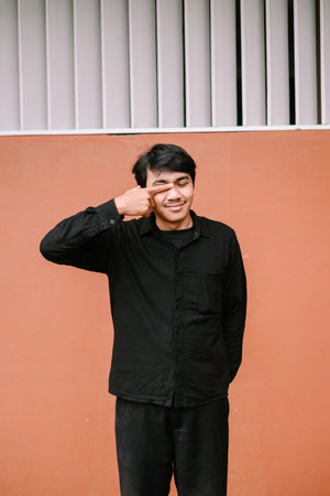 A happy young man in a black shirt makes a playful eye gesture with closed eyes and a smile while...