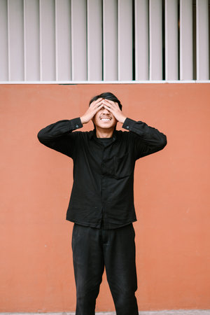 A young man stands against an orange wall, covering his eyes with his hands while showing a...