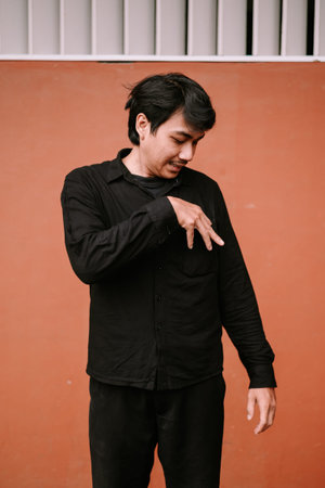 A young Asian man wearing a black shirt stands thoughtfully against a vibrant orange wall, making an...