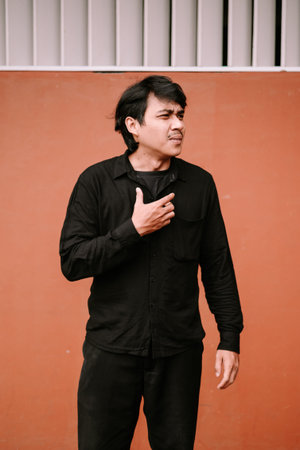 A young man with a thoughtful and slightly confused expression places his hand on his chest. This...