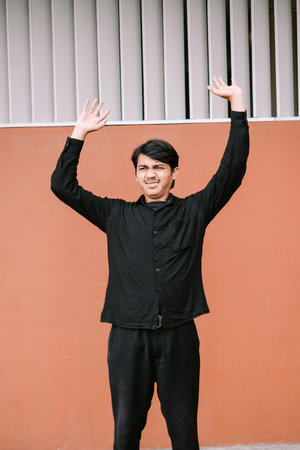 A young Asian man stands against an orange wall, raising both hands in a gesture of frustration and...