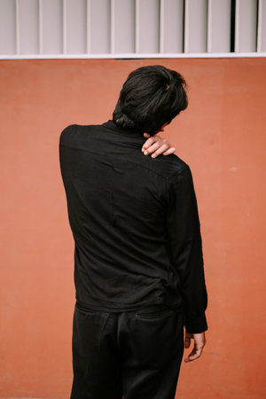 A person, seen from the back, gently touches their neck and shoulder, possibly experiencing pain,...