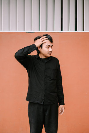 A thoughtful young man in a black shirt poses with his hand on his head against an orange-brown...