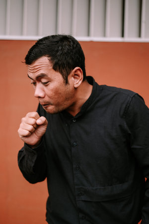 An adult man covers his mouth with his fist, preventing the spread of germs while coughing. This...