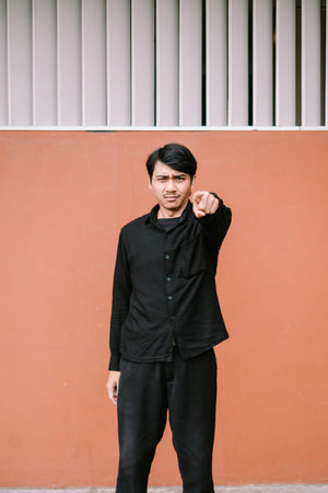 A serious young man in black attire points his finger directly at the camera, standing against an...