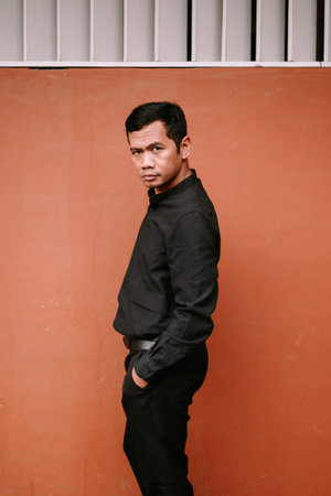 A thoughtful man in a black collared shirt and dark pants stands with hands in pockets against a rustic orange wall, looking directly at the camera with a serious expression.の写真素材