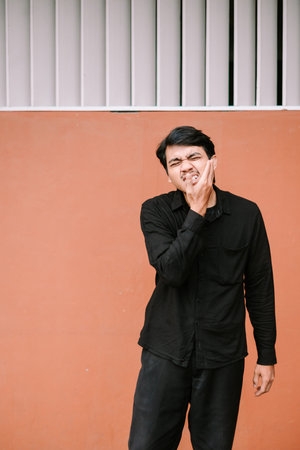 A young man is grimacing and holding his cheek due to intense toothache or jaw pain, standing...