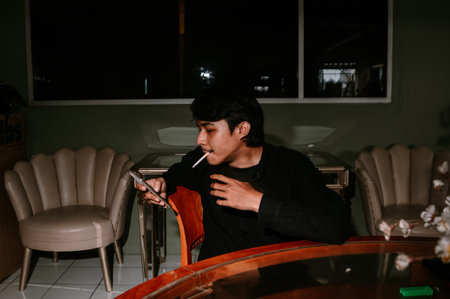 A young man, casually dressed, takes a moment to relax, smoking a cigarette and checking his...