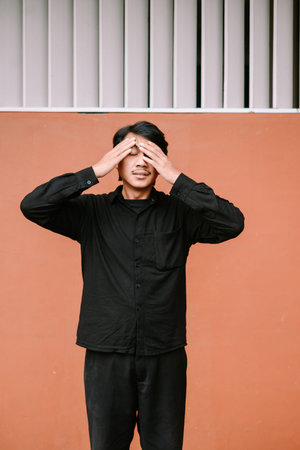 A young man covers his eyes with his hands, displaying feelings of stress, fatigue, or discomfort....