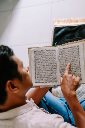 A man focuses on reading the Holy Quran, his finger tracing the sacred Arabic verses. He is engaged...