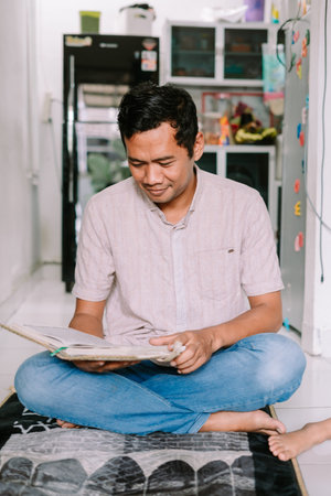 An adult man sits on a prayer mat, deeply focused on reading a holy book at home. He is dressed...