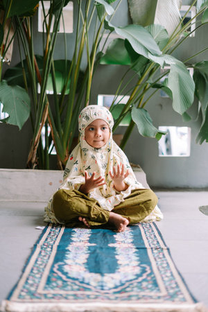 A young Muslim girl, wearing a floral mukena, sits cross-legged on a traditional blue prayer mat....