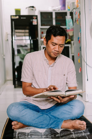 An adult man sits cross-legged on a mat, intently reading from a book. He is focused on his reading...